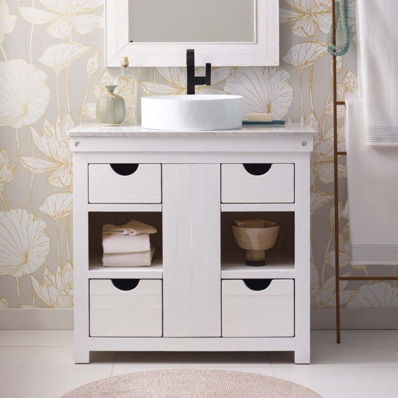 A wooden freestanding cabinet has four drawers and two alcoves with a circular sink on top. Behind is a floral wallpaper and a square mirror and to the right hand side a ladder style towel rack. 