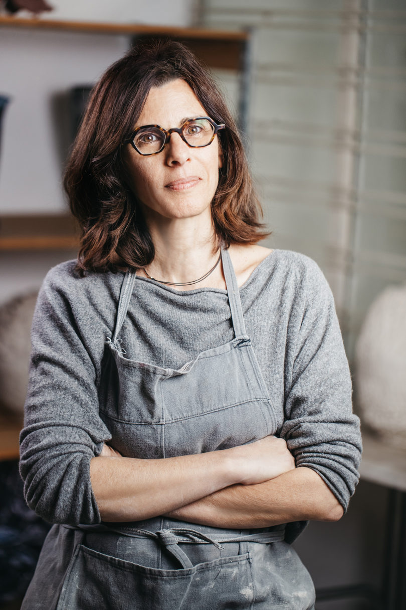 light-skinned woman with long brown hair wearing glasses and a grey shirt and work apron