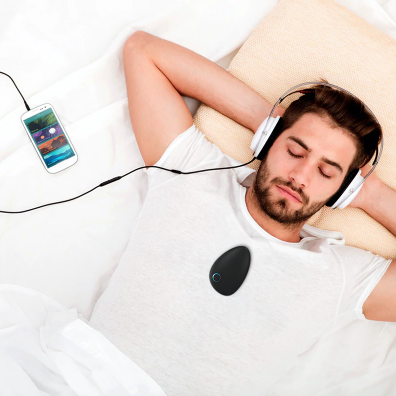 light-skinned man with dark hair and facial hair lays with his hands behind his head, wearing headphones connected to his smartphone with a pebble-shaped device resting on his chest