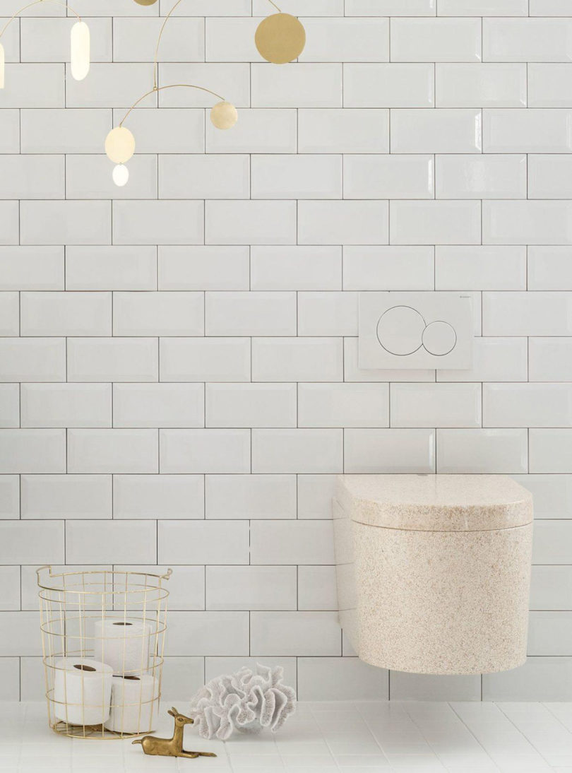 Neutral natural hue bathroom with Block wall hung toilet, brass colored wire basket with four rolls of toilet paper and a hanging mobile of circles dangling from the ceiling.