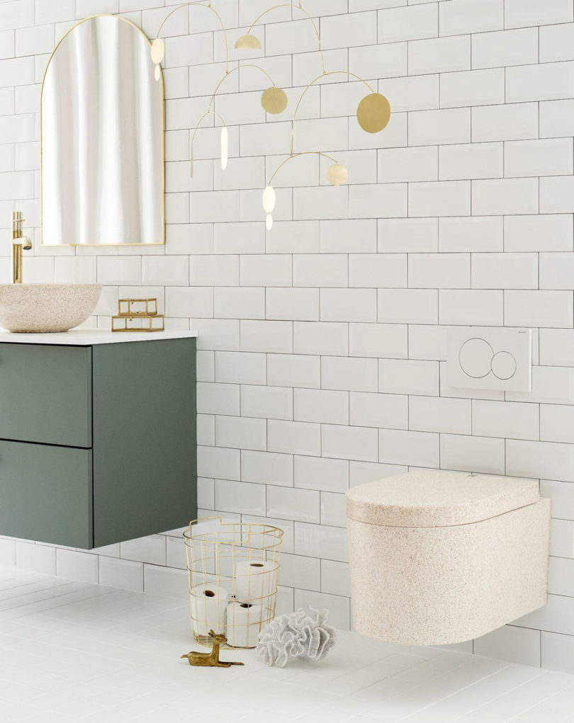 Neutral natural hue bathroom with freestanding on-top counter basin sink with arched mirror and green cabinetry with Block toilet on the right with nearby brass colored wire basket with four rolls of toilet paper and a hanging mobile of circles dangling from the ceiling.