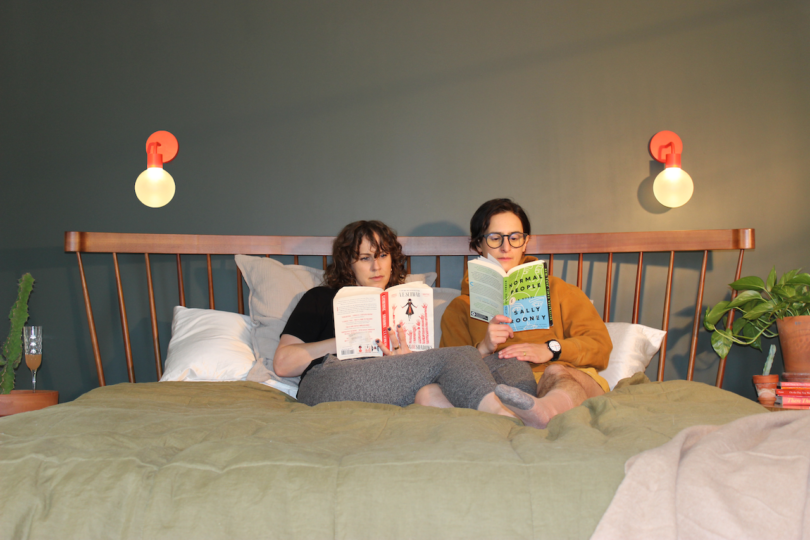 two women sitting in bed next to two red wall lights