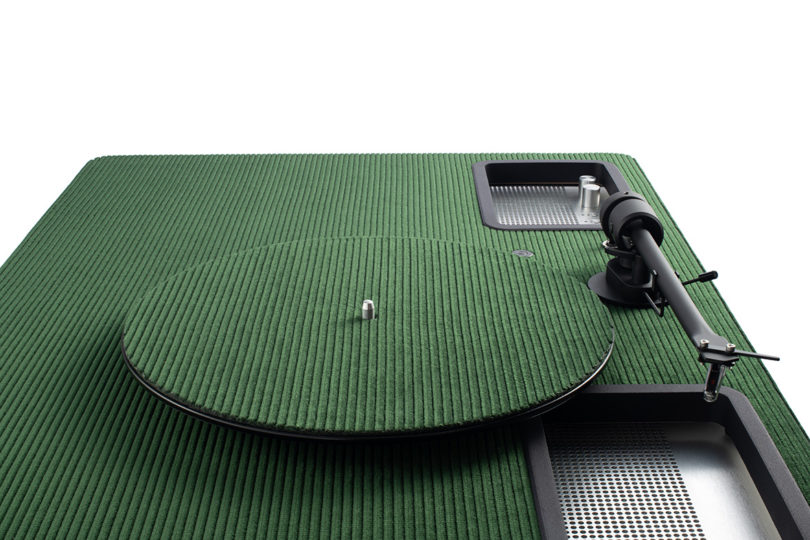 Green Riga corduroy velvet topped turntable and speaker system set in room surrounded by curtain backdrops from overhead side view showing turntable arm and platter.