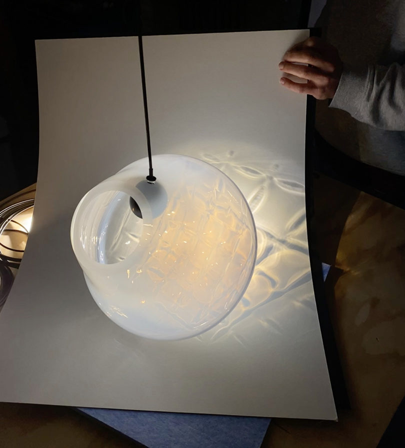 Person holding a large piece of white paper in front of opaque white GRID handblown glass with spherical LED spotlight to show the unique patterns seen passing through the textured glass surface.
