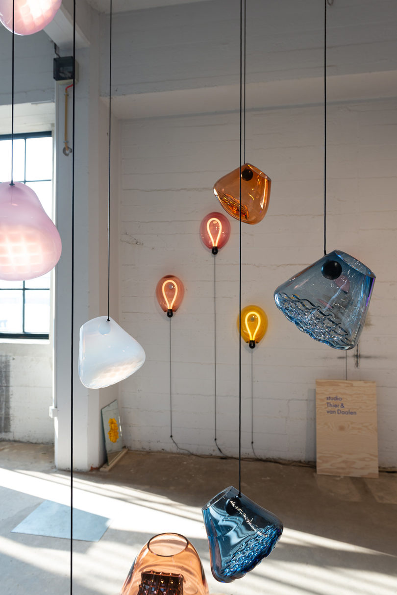 Six hanging GRID pendant glass lights in orange, opaque pink, white and light blue BULLA wall lights in background on white brick wall.