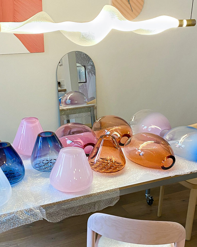 An array of handblown glass GRID pendants and BULLA wall lights in various colors displayed across bubble wrap covered table with wavy ceiling light above and arched mirror in background.