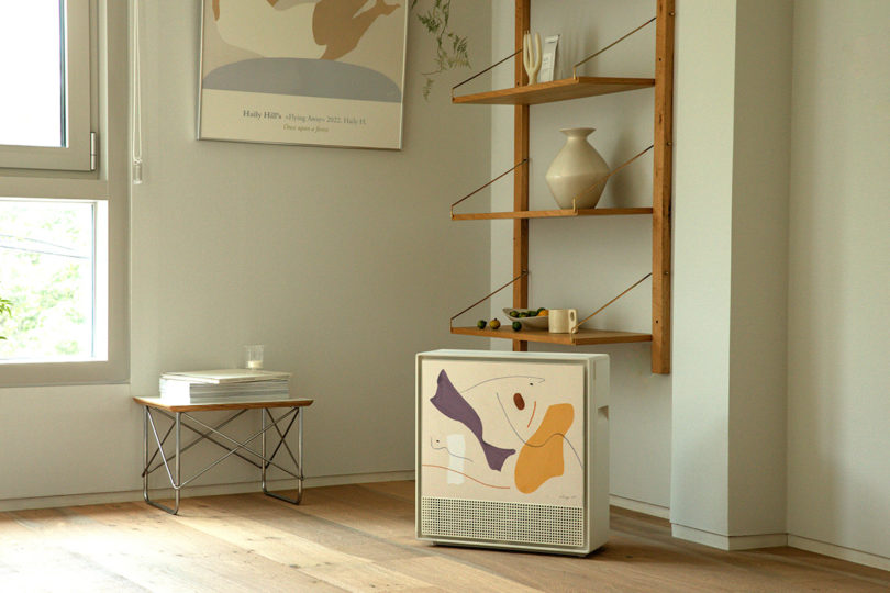Coway Airmega 250 Art on ground near wall mounted bookshelves with small ceramic pieces set on it and small table with magazines and candle set on top.