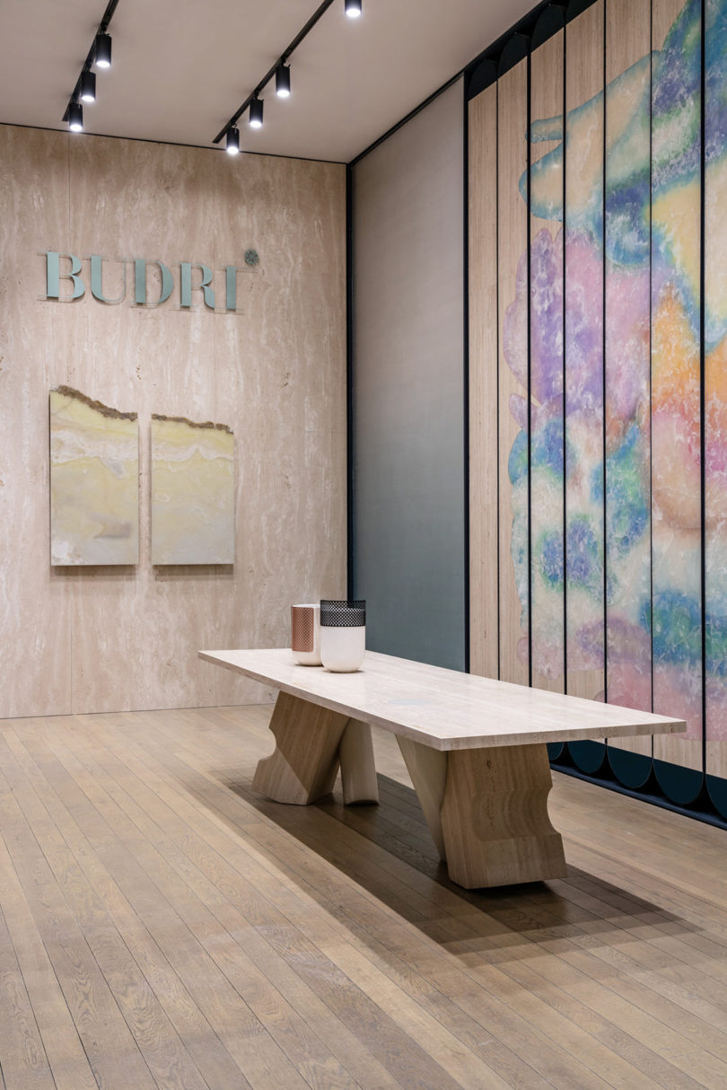 exhibit entrance with signage reading "BUDRI", a long dining table, and a wall installation resembling a watercolor painting