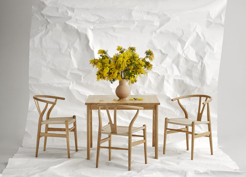 three wishbone chairs in oak next to a dining table with a vase of yellow flowers