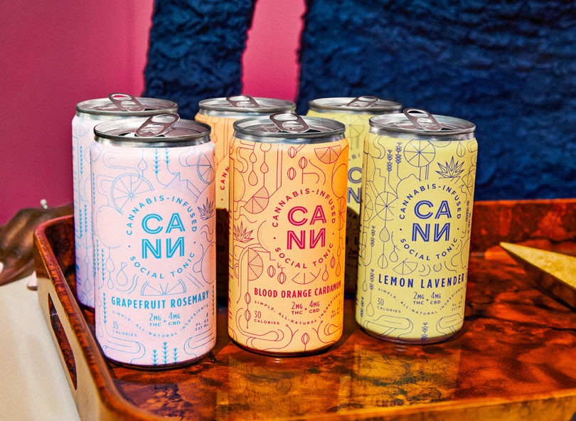 Six cans of Cann cannabis-infused beverages with grapefruit, blood orange, and lemon lavender flavors on wooden tray with person's torso in background. 