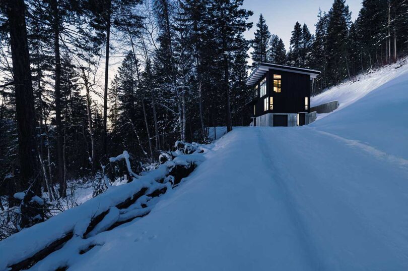evening view of exterior of modern black cabin surrounded by snow