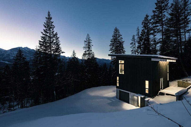 evening view of exterior of modern black cabin surrounded by snow
