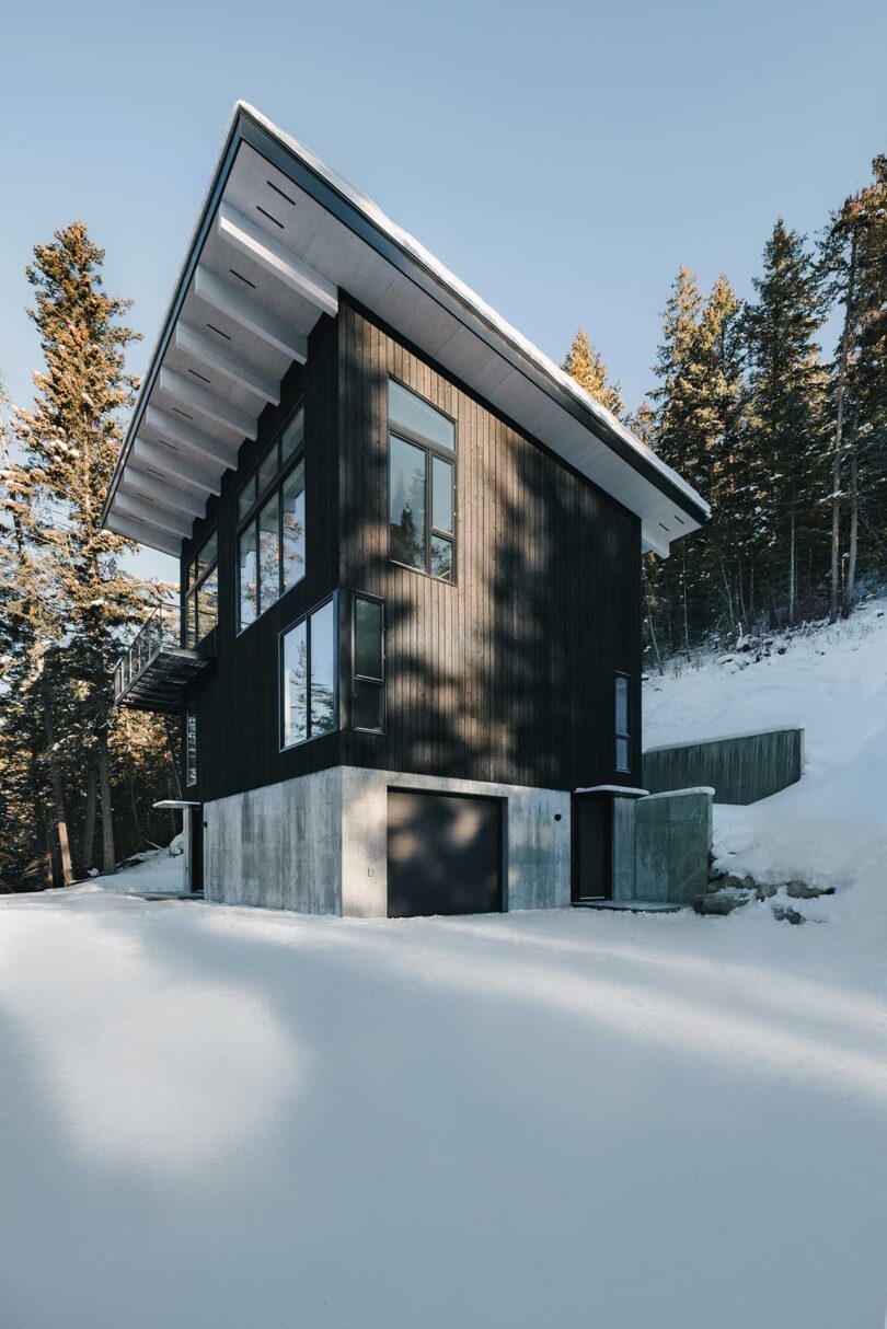 angled exterior view of modern black and white cabin with concrete base surrounded by snowy hills and trees