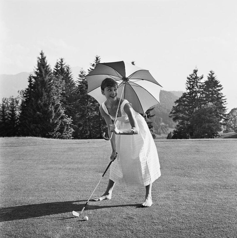 black and white image of Audrey Hepburn golfing while holding an umbrella