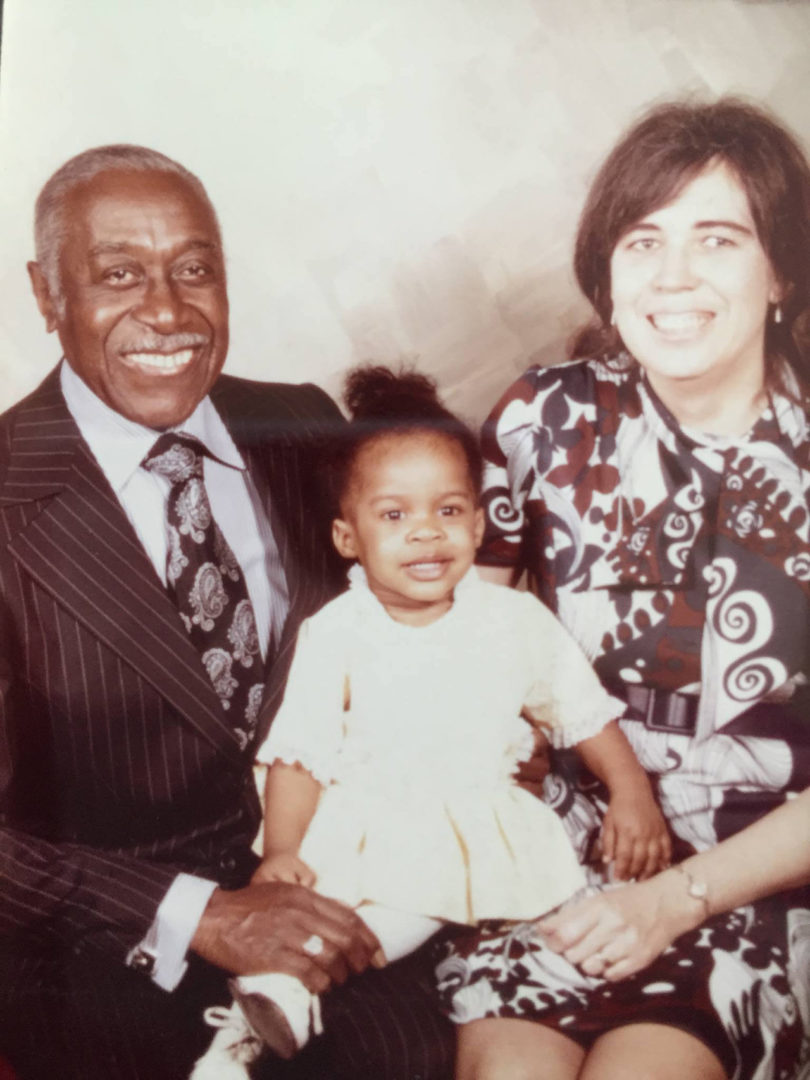 brown-skinned man wearing a suit, light-skinned woman with dark hair wearing a patterned dress, and a brown-skinned baby girl in a white dress posing for a family portrait