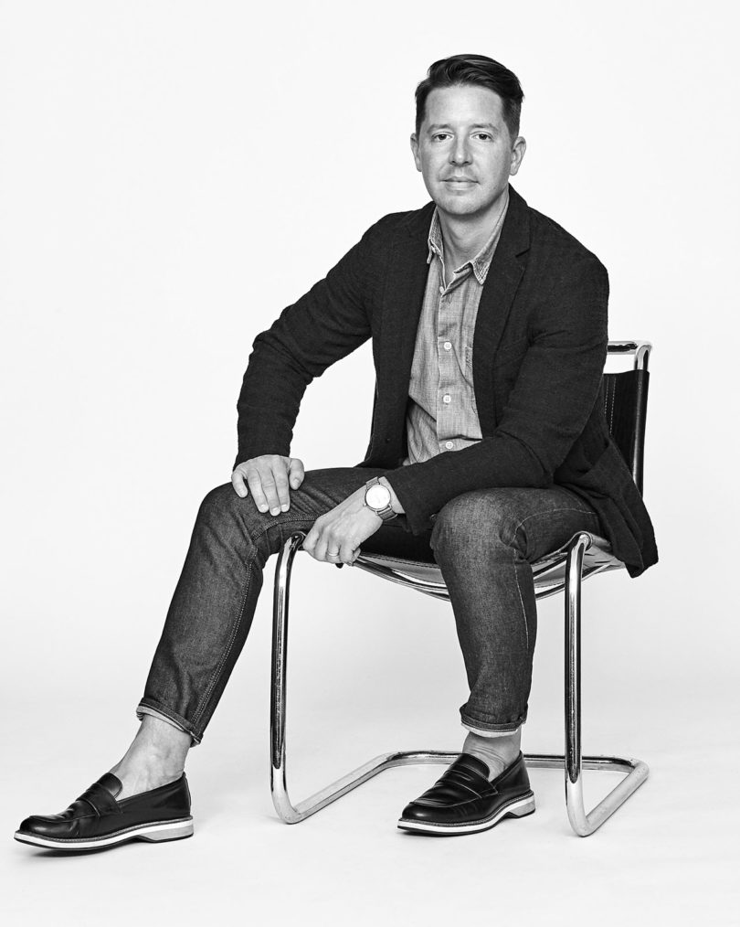 black and white image of a man sitting in a chair wearing jeans, a button down shirt, blazer, and loafers