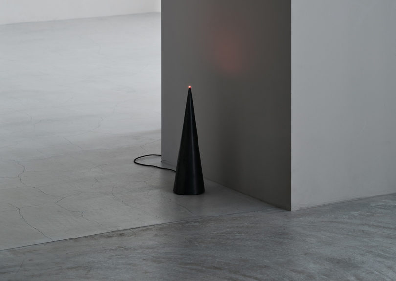 Flight, an all black cone shaped floor or table lamp with a single bright red LED light at the tip set on concrete floor near wall with soft red reflection on gray walls.