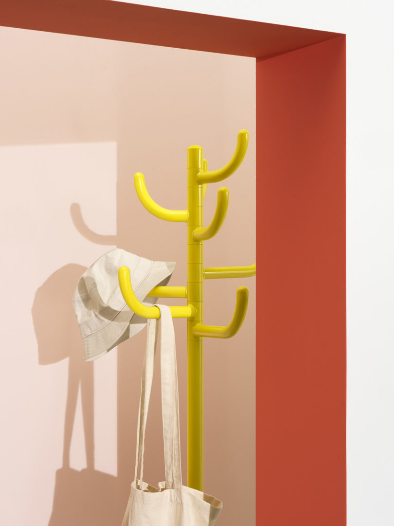 Bright yellow tubular coat rack in red and peach painted wall room corner with hat and tote bag hanging from the rack arms.