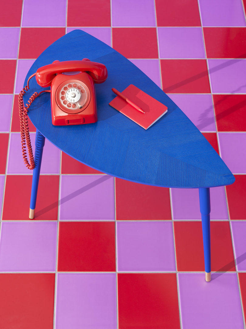 Red rotary phone on bright blue three-legged side table placed on red and violet checkerboard tile flooring.