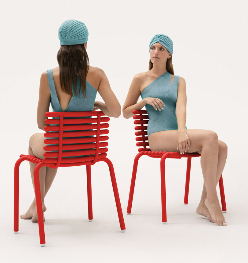 two light-skinned woman with long dark hair wearing grey-blue one-shoulder swimsuits and matching swimming caps sit opposite one another in red outdoor chairs
