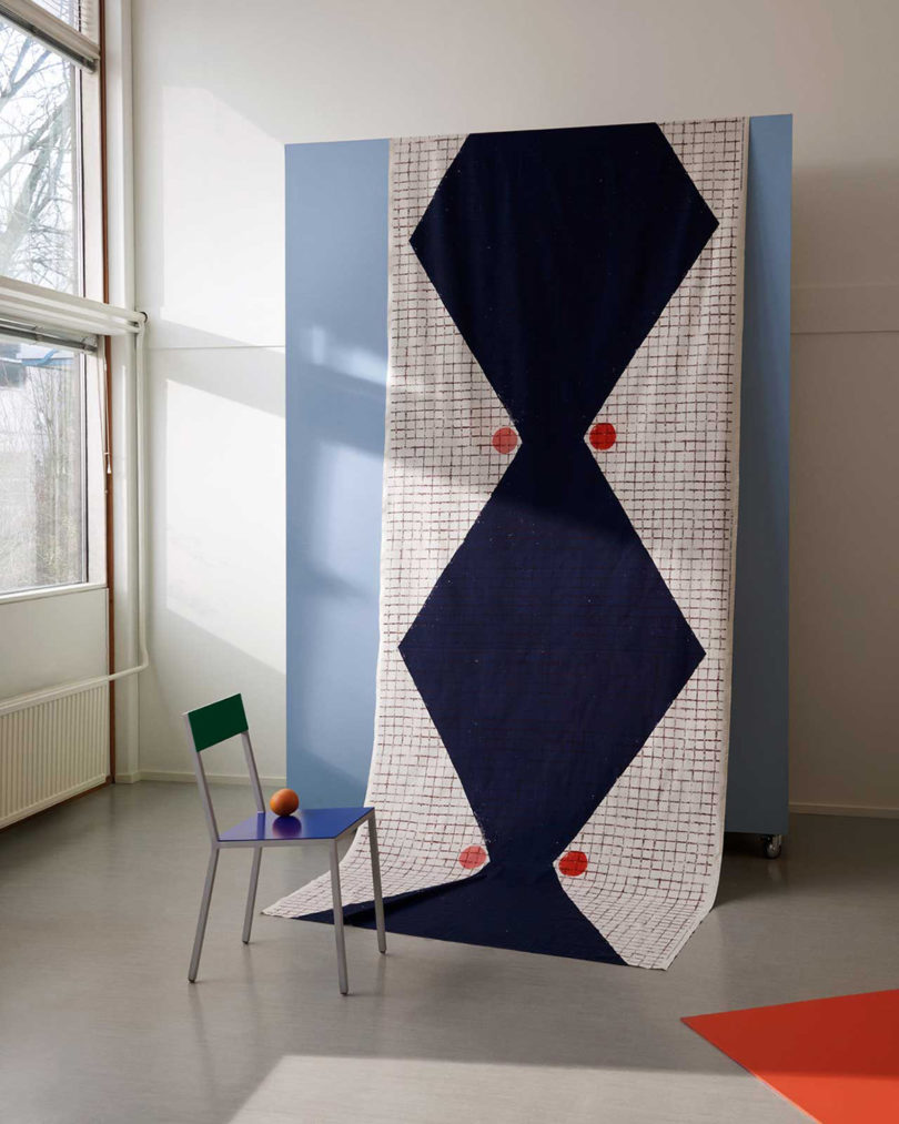 Large Marimekko x Finkenauer Artist Series textile draped over blue wall with blue metal chair and window nearby