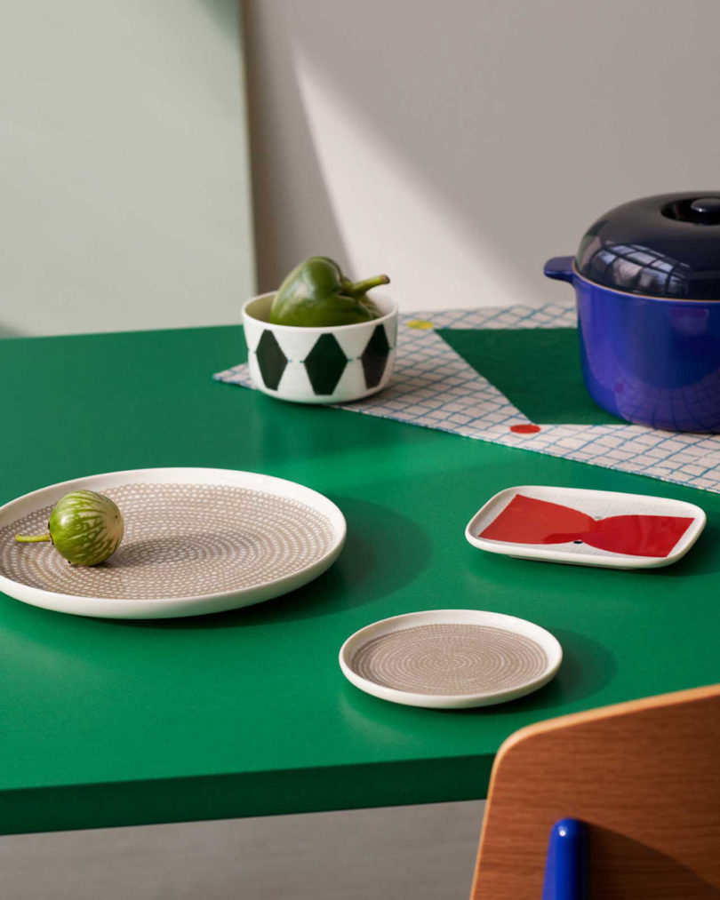 Green table top with ceramic plates, tray, and bowl with rich blue cast iron pot set on Marimekko fabric.