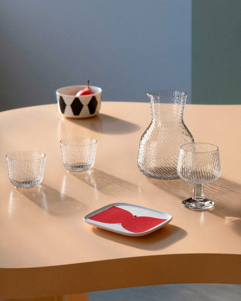 مجموعه کاسه و سینی مجموعه Marimekko x Finkenauer در کنار مجموعه ظروف شیشه ای Matti Klenell روی میز گرد هلویی مایل به قهوه ای با زمینه دیوار آبی.
