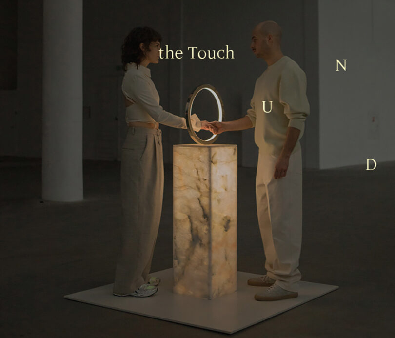 A woman and man shaking hands through a large ring with illuminated center on top of a rectangular pedestal with the words "the Touch" printed over the photo.