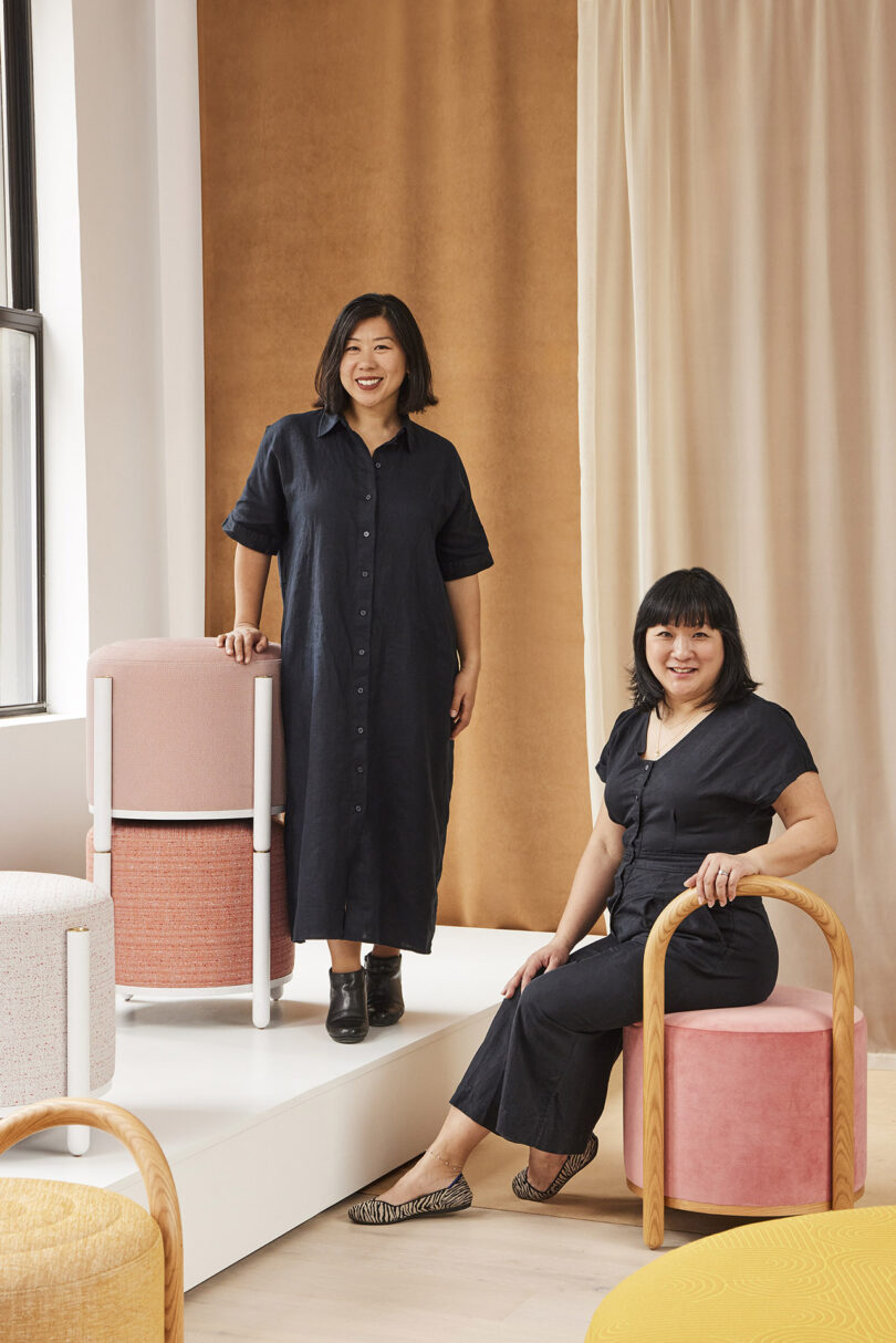 two women with dark hair, wearing black dresses and posing with pink pouf stools for a portrait