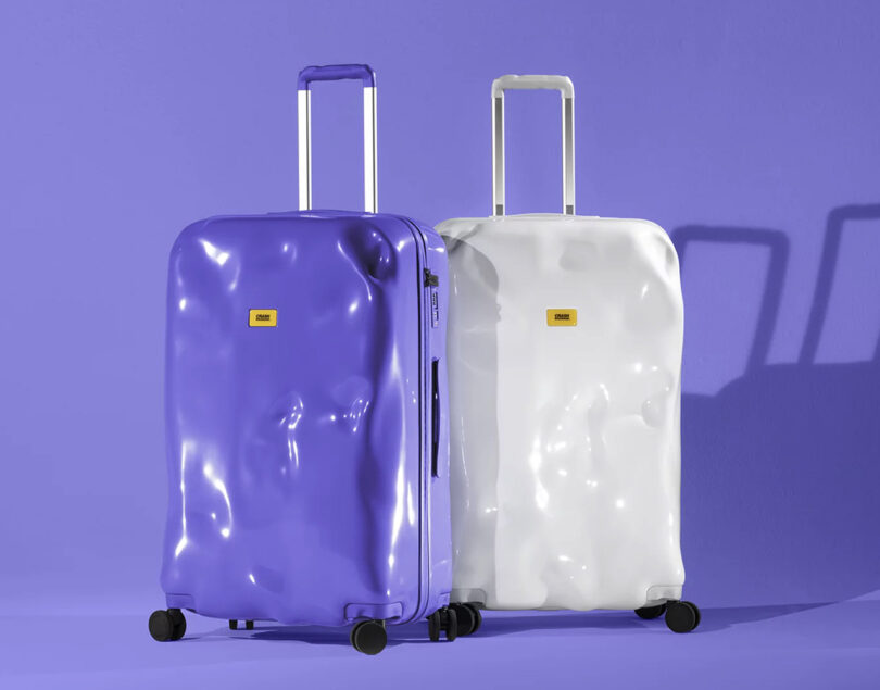 A pair of Crash Baggage roller suitcases in purple and white in a purple background room.