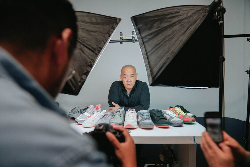 light-skinned bald man wearing a black shirt sits behind a table displaying shoes