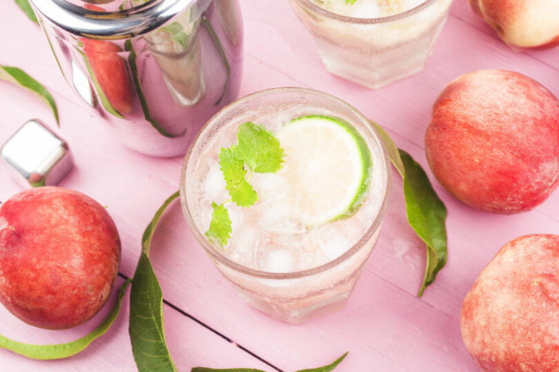 overhead photo of a cocktail with a lime surrounded by peaches on a pink background
