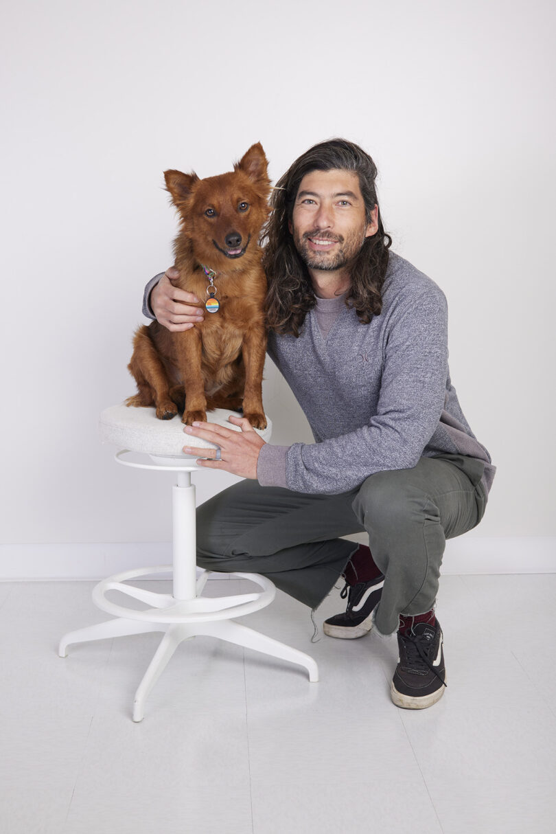 brown-skinned man with long dark hair posing with a copper colored dog who sits on a stool