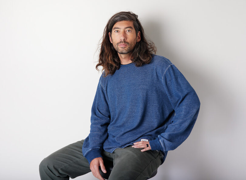 brown-skinned man with long dark hair wearing a blue sweatshirt and dark pants sits and looks into the camera