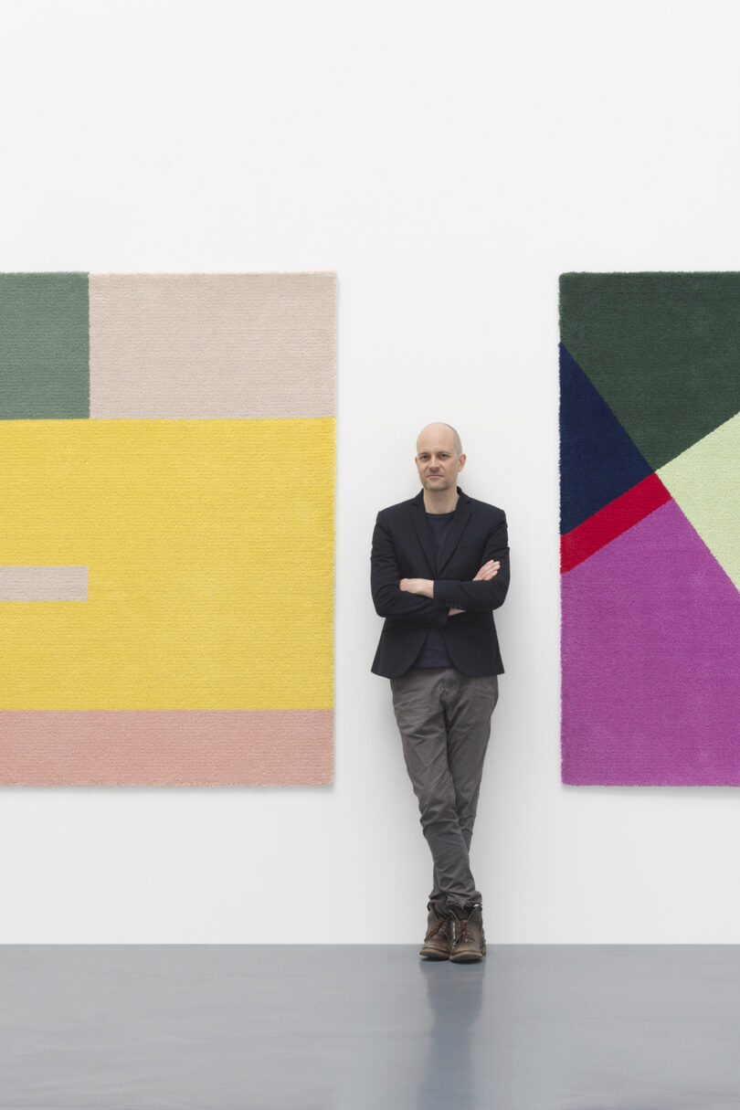 two colorful geometric rugs hanging on a white gallery wall with a light-skinned man standing for a portrait in between them