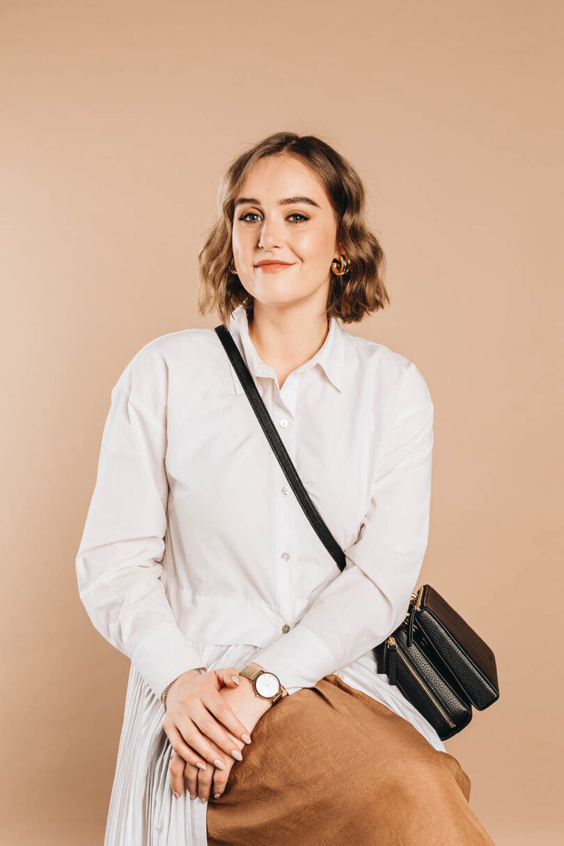 light-skinned woman with short brown hair wearing a button-down white shirt and a crossbody black bag