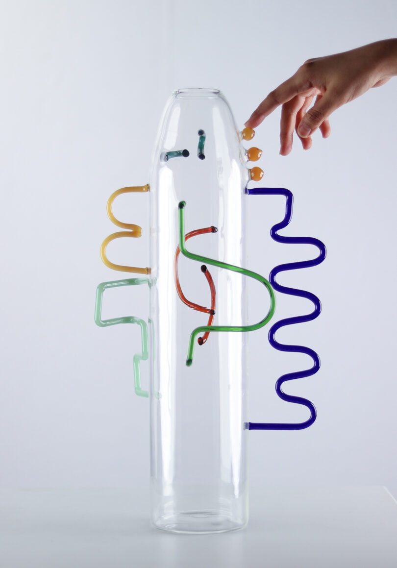 a light-skinned hand touching a clear glass cylinder with abstract colored shapes of glass attached to the exterior