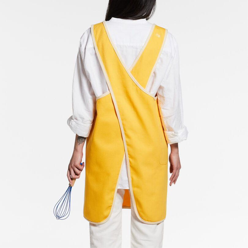 the back of a person wearing a bright yellow kitchen apron and holding a whisk