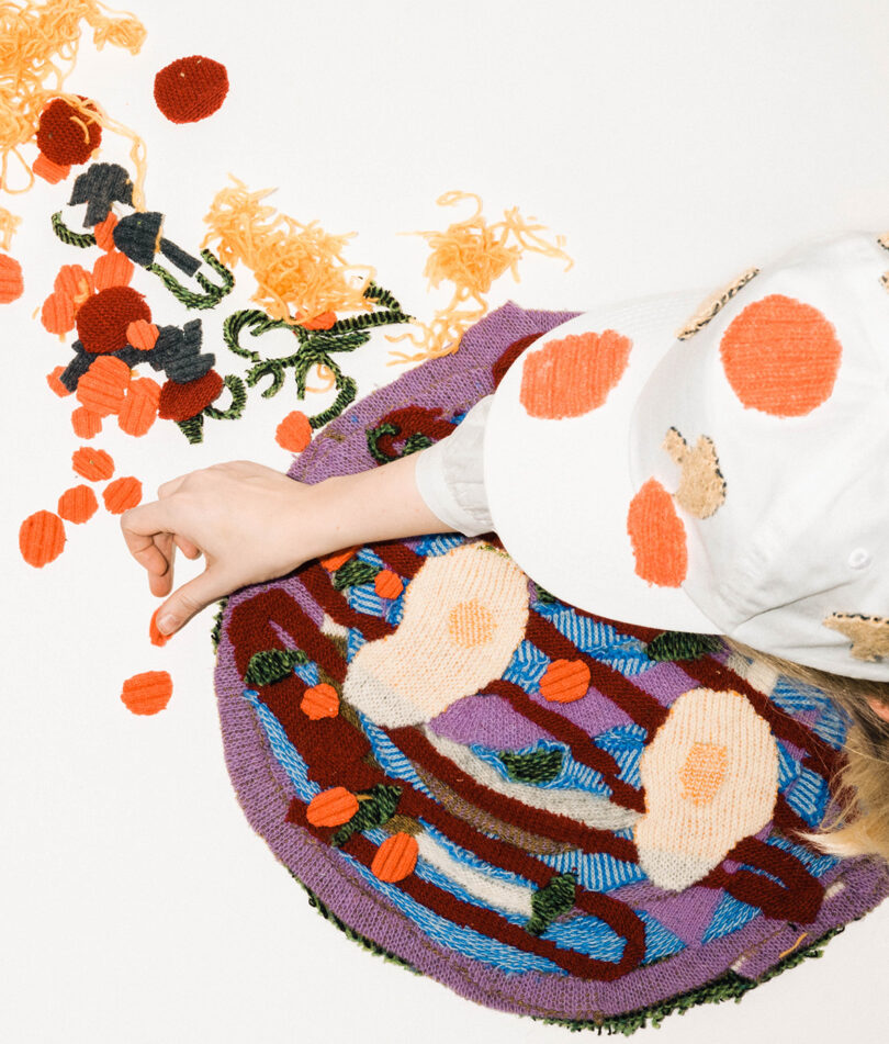 Woman in white baseball cap and t-shirt topping a fake pizza made from wool remnants.