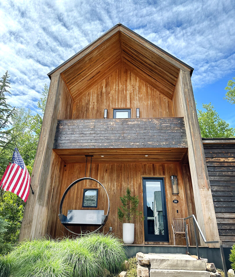 خانه مدرنیستی با روکش چوبی با پرچم آمریکا در بیرون و نیمکت چرخان دایره‌ای بزرگ در جلو.
