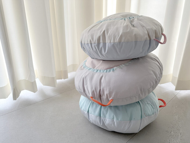 Stack of three round pastel colored pillows made from recycled car air bags with orange handles with curtains in background.