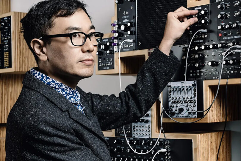 Bespectacled Yuri Suzuki of Pentragram posing with Moog synthesizers, in the act of plugging into ports.