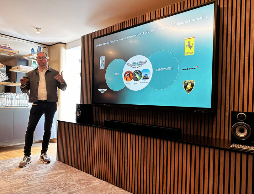 Aston Martin director of design Miles Nurnberger to the side of large flat screen television showing graphics about the brand's positioning landing between the luxury and performance automotive categories.