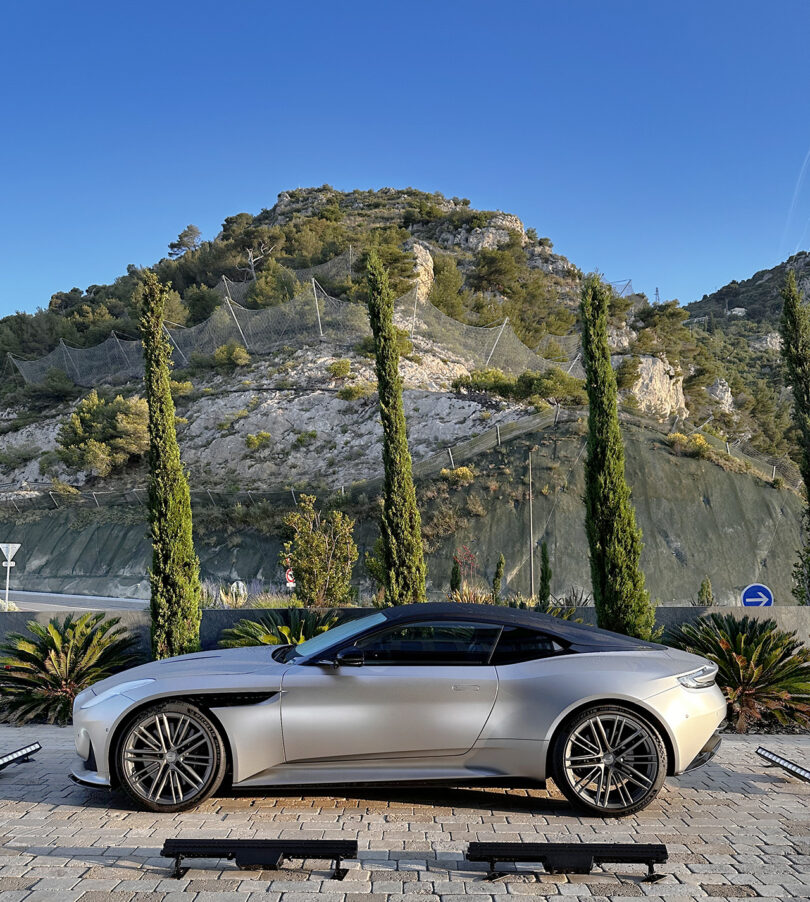 Side shot of parked Silver Aston Martin parked at Cote d'Azur hotel parking area with mountainside in near background and three Cypress trees.