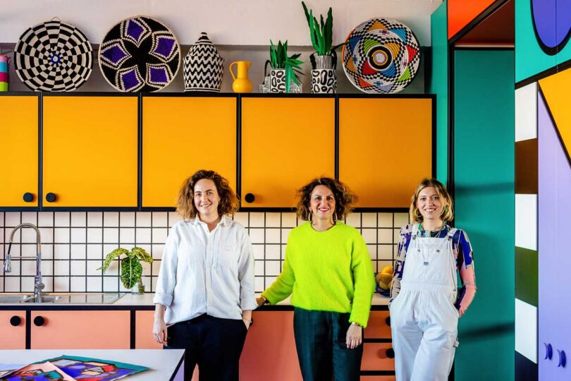 three women standing in front of colorful kitchen
