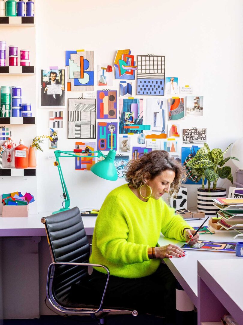 artist sitting sideways in bright yellow sweater at desk surrounded by art