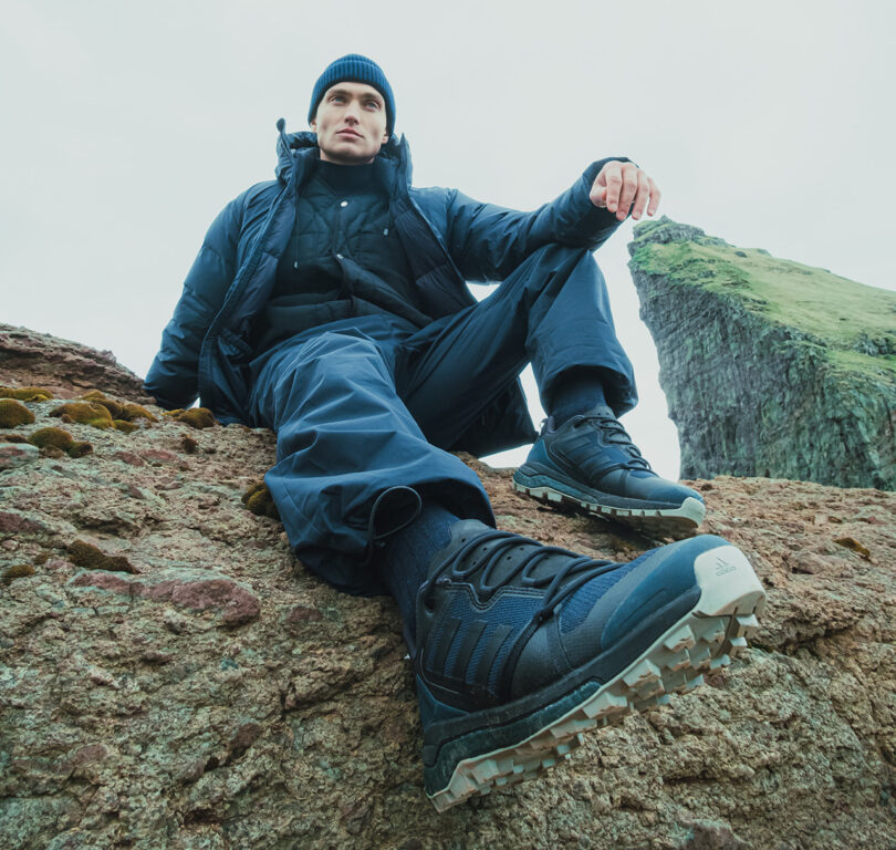 Young man in beanie and puffy jacket seated on a jutting piece of the Faroe Islands with his footwear prominently in the foreground.