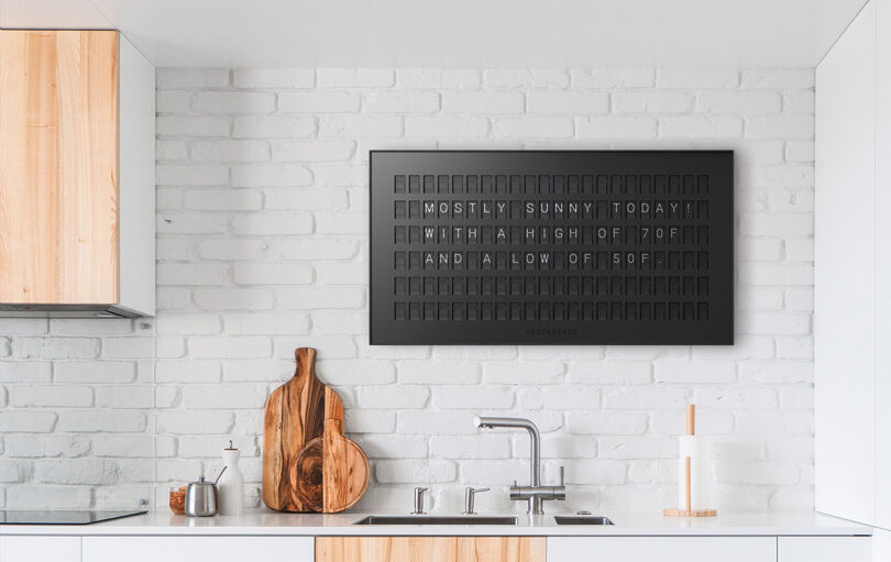 Vestaboard in a chic matte black wall mounted onto a white painted brick wall in a kitchen.