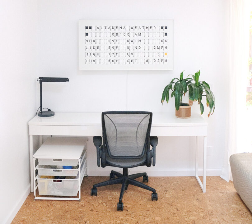 White narrow desk on cork floors, with Humanscale office chair, black table lamp, small house plant, and wall mounted Vestaboard in white displaying weather forecast.