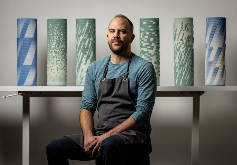 Artist and designer Brian Peters seated wearing apron in front of 6 of his 3D printed sculptures.